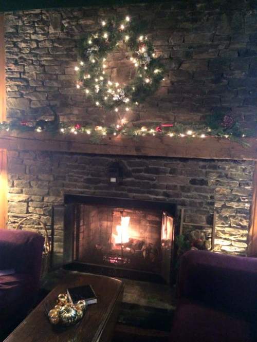 Cozy living room with a stone fireplace, decorated with a wreath and lights, glowing warmly with a fire.