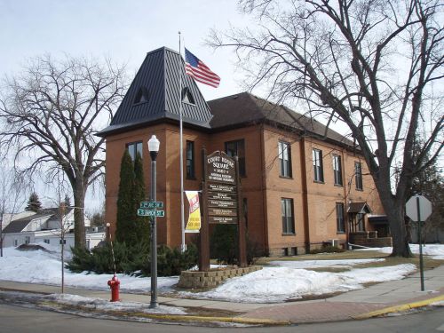 Historic brick building with a peaked roof, American flag, and sign, surrounded by snow and trees.