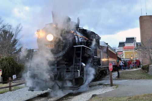 A vintage steam train billows smoke as it approaches a station, with people gathered nearby.