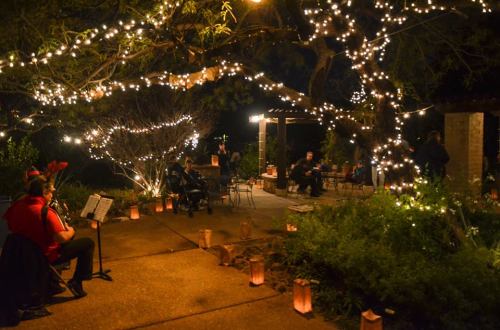 A cozy outdoor scene at night, with string lights, lanterns, and people enjoying music and conversation.