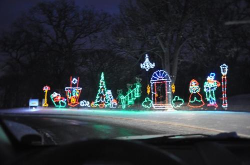 Colorful holiday lights decorate a snowy roadside, featuring trees, gifts, and festive characters against a dark sky.