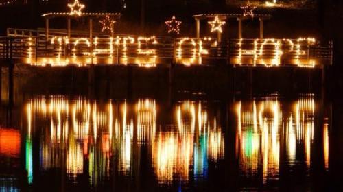 "Illuminated sign reading 'Lights of Hope' reflected in water, surrounded by festive decorations and stars."