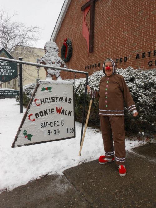 A person dressed as a gingerbread man stands next to a snowy sign for a Christmas cookie walk event.