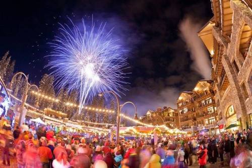 A festive night scene with a crowd enjoying a fireworks display over a holiday market, surrounded by twinkling lights.
