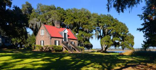 A charming brick house with a red roof, surrounded by lush greenery and large trees, near a calm water body.