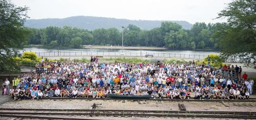 A large group of people gathered outdoors near a river, with trees and mountains in the background.