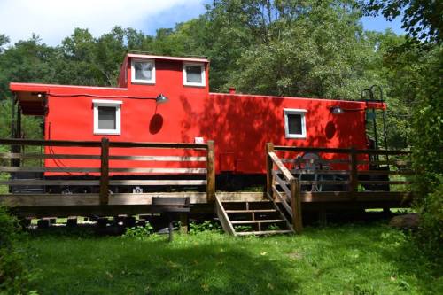 A bright red caboose on a wooden platform surrounded by trees and grass, featuring steps and outdoor seating.