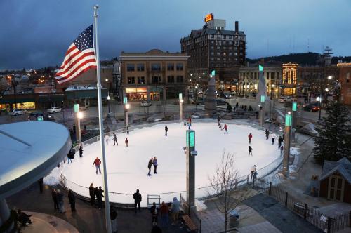 A winter scene of an outdoor ice skating rink with skaters, surrounded by buildings and an American flag waving.