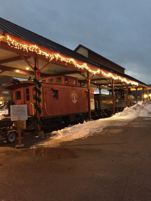 A red train caboose decorated with holiday lights, under a covered platform with snow on the ground.