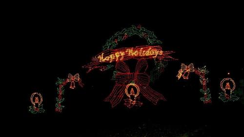 Colorful holiday lights display featuring a "Happy Holidays" sign, bells, and candles against a dark background.