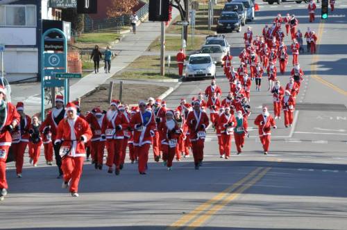 A large group of runners dressed as Santa Claus races down a city street during a festive event.