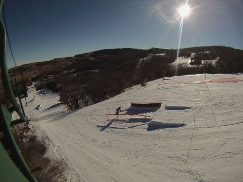 A skier performs a trick on a snowy slope, with a sunlit landscape and ski lift in the background.