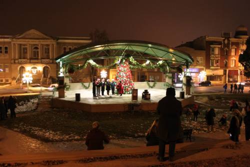 A festive outdoor stage with a Christmas tree, people gathered, and holiday decorations at night.