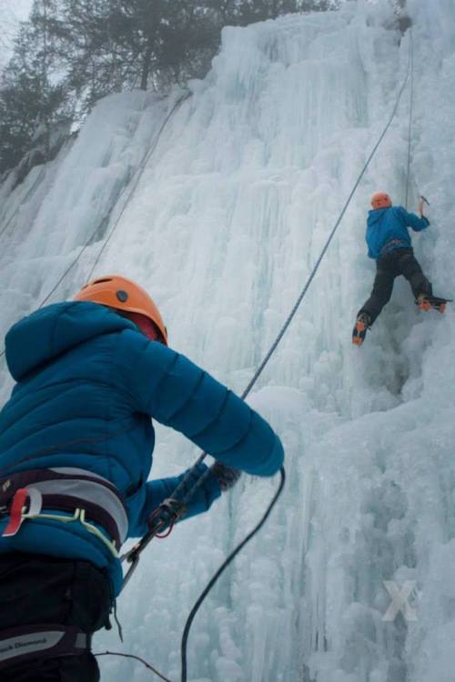 Two climbers ascend a frozen ice wall, wearing helmets and climbing gear, with ropes secured for safety.