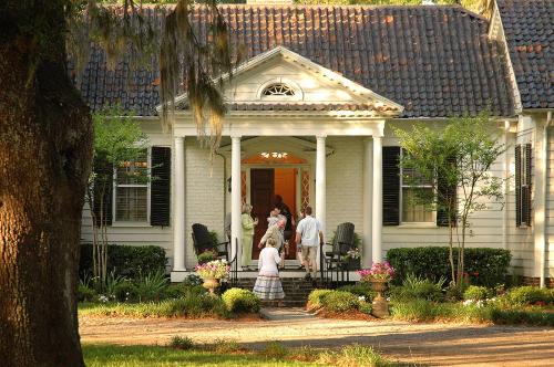 A charming white house with a porch, surrounded by greenery, as people walk towards the entrance.