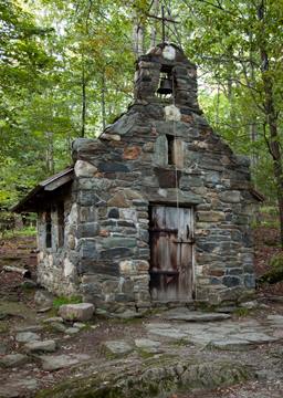 A small, weathered stone chapel with a bell tower, surrounded by lush green trees in a forested area.