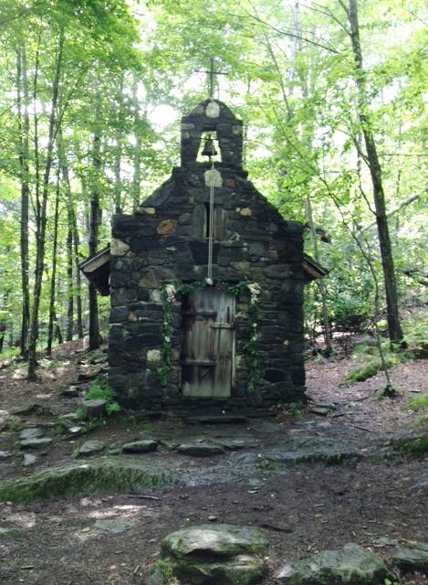 A small stone chapel surrounded by trees, featuring a wooden door and a bell on top, set in a forested area.