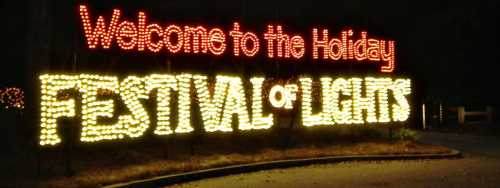 Brightly lit sign reading "Welcome to the Holiday Festival of Lights" at night.