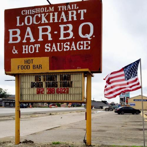 Sign for Chisholm Trail Lockhart Bar-B-Q & Hot Sausage, featuring a message about ordering turkey, with an American flag nearby.