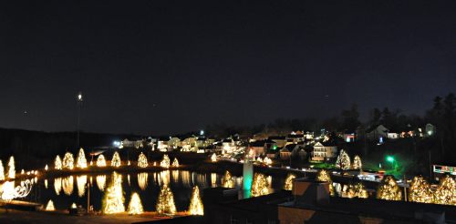 A nighttime view of a festive town with illuminated trees and buildings reflecting in a calm lake.