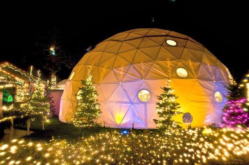 A geodesic dome illuminated at night, surrounded by colorful Christmas lights and decorated trees.