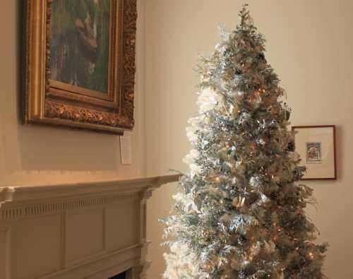 A decorated Christmas tree stands beside a fireplace and a framed painting on the wall in a cozy room.