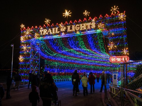 A colorful archway of lights reading "Trail of Lights" with people walking underneath at night.