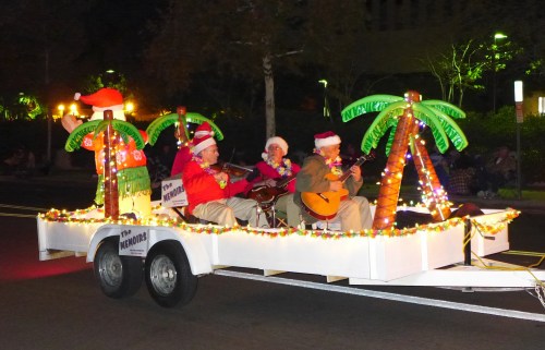 A festive parade float with musicians in Santa hats, surrounded by palm trees and colorful lights.