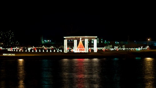 A festive display of Christmas lights around a gazebo by the water, reflecting on the surface at night.