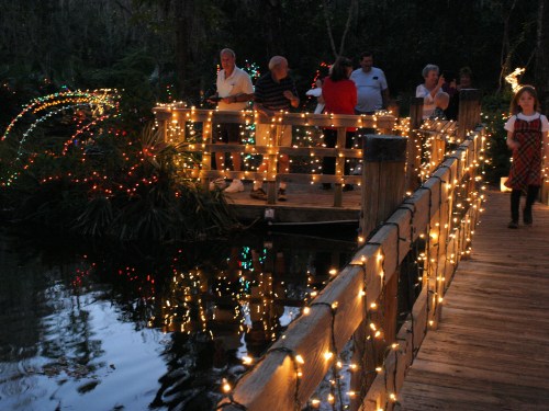 A wooden bridge adorned with colorful lights, with people enjoying the festive atmosphere by a calm waterway.