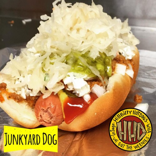 A hot dog topped with chili, onions, sauerkraut, and pickles in a bun, with a logo for Hillbilly Hotdogs.