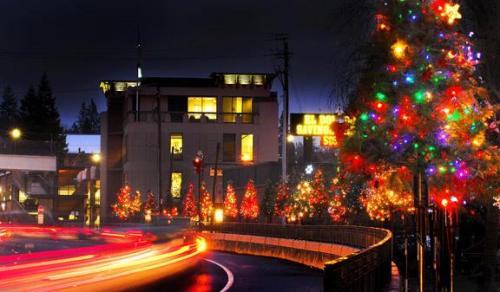 Colorful Christmas trees line a street at night, with festive lights and blurred car lights creating a cheerful atmosphere.