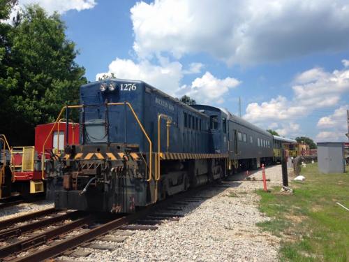 A blue train engine with a passenger car parked on a gravel track under a partly cloudy sky.