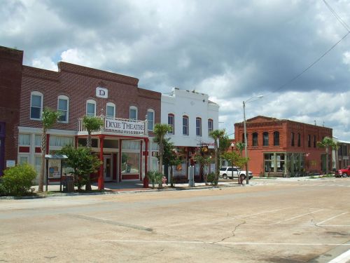 Historic buildings line a street, featuring the Dixie Theatre with palm trees and a cloudy sky above.