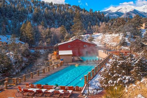 A scenic outdoor hot spring pool surrounded by snow-covered mountains and trees, with lounge chairs nearby.