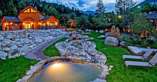 A cozy cabin surrounded by mountains, featuring a stone pathway, a pond, and lounge chairs in a serene outdoor setting.