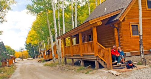 A couple and a dog sit on the porch of a log cabin, surrounded by trees and a dirt road in a scenic, autumn setting.