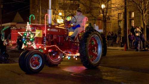 A decorated red tractor with lights, driven by a person in winter attire, parades through a festive street at night.