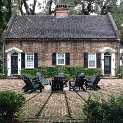 A charming brick house with a slate roof, flanked by green shrubs and a circular seating area of black chairs.