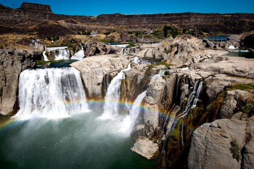 A scenic view of waterfalls with a rainbow arching over the water, surrounded by rocky terrain and greenery.