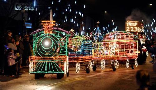 A brightly lit parade float shaped like a train, adorned with colorful lights, moves through a festive street.