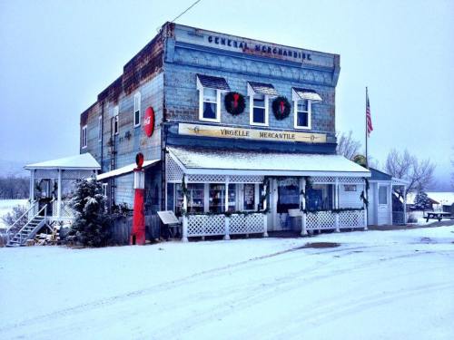 A rustic general store covered in snow, adorned with wreaths, with an American flag nearby. Winter scene.