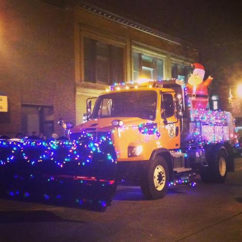 A decorated snowplow truck with colorful lights and a Santa figure, parked in a festive nighttime setting.