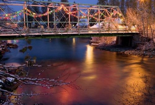 A snowy bridge adorned with colorful lights spans a calm river, reflecting warm hues in the evening twilight.