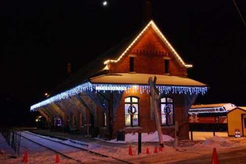 A charming train station decorated with festive lights, surrounded by snow, under a night sky with a crescent moon.
