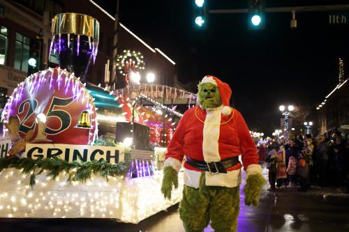 The Grinch in a Santa suit walks beside a festive parade float decorated with lights and holiday themes.