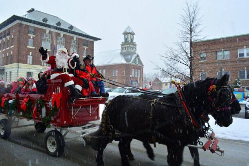A festive horse-drawn sleigh with Santa and children, surrounded by snow, passing a historic building in winter.