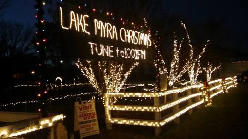 Sign reading "Lake Myra Christmas" with festive lights and decorations in a nighttime setting.