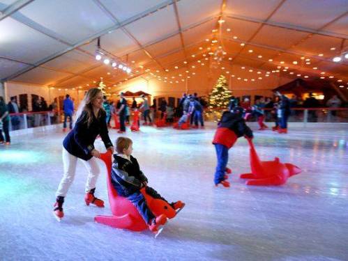 A woman pushes a child on a penguin-shaped sled on an ice rink, surrounded by festive decorations and other skaters.