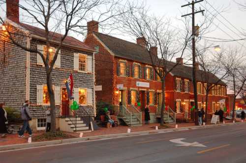 Historic buildings decorated with holiday lights, people walking along the street, and bare trees in the evening.